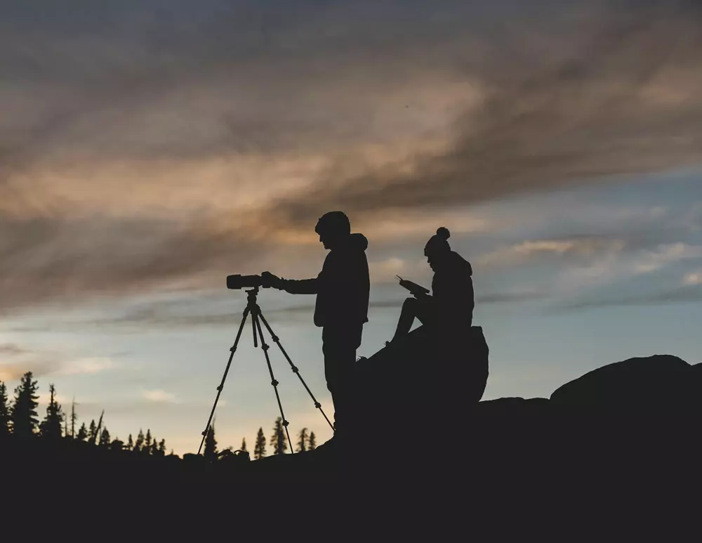 Silhouette of two photographers on a mountain peak, capturing the sunset over the horizon.
